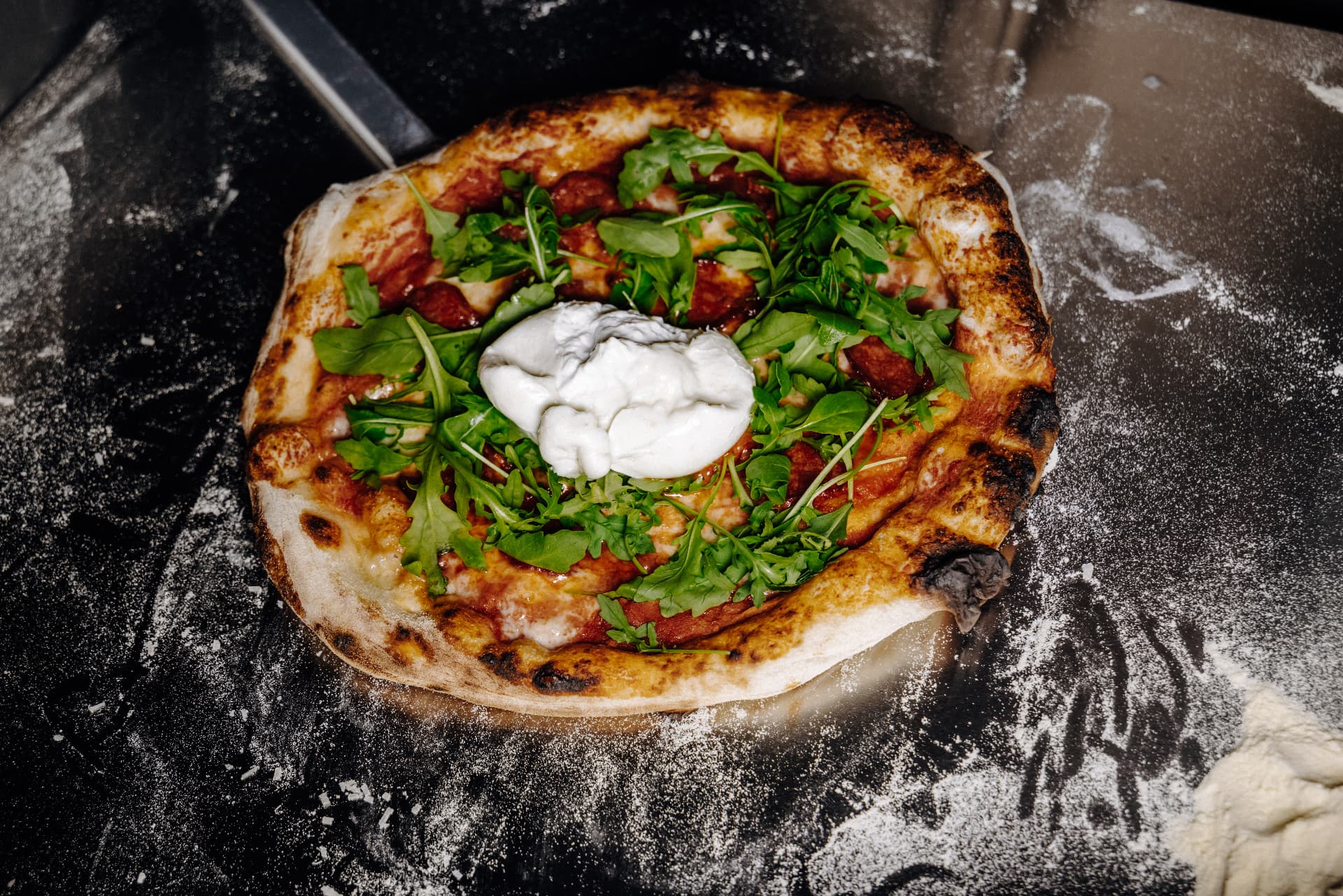 Frisch gebackene Pizza mit Rucola und Burrata auf einer dunklen bemehlten Arbeitsfläche