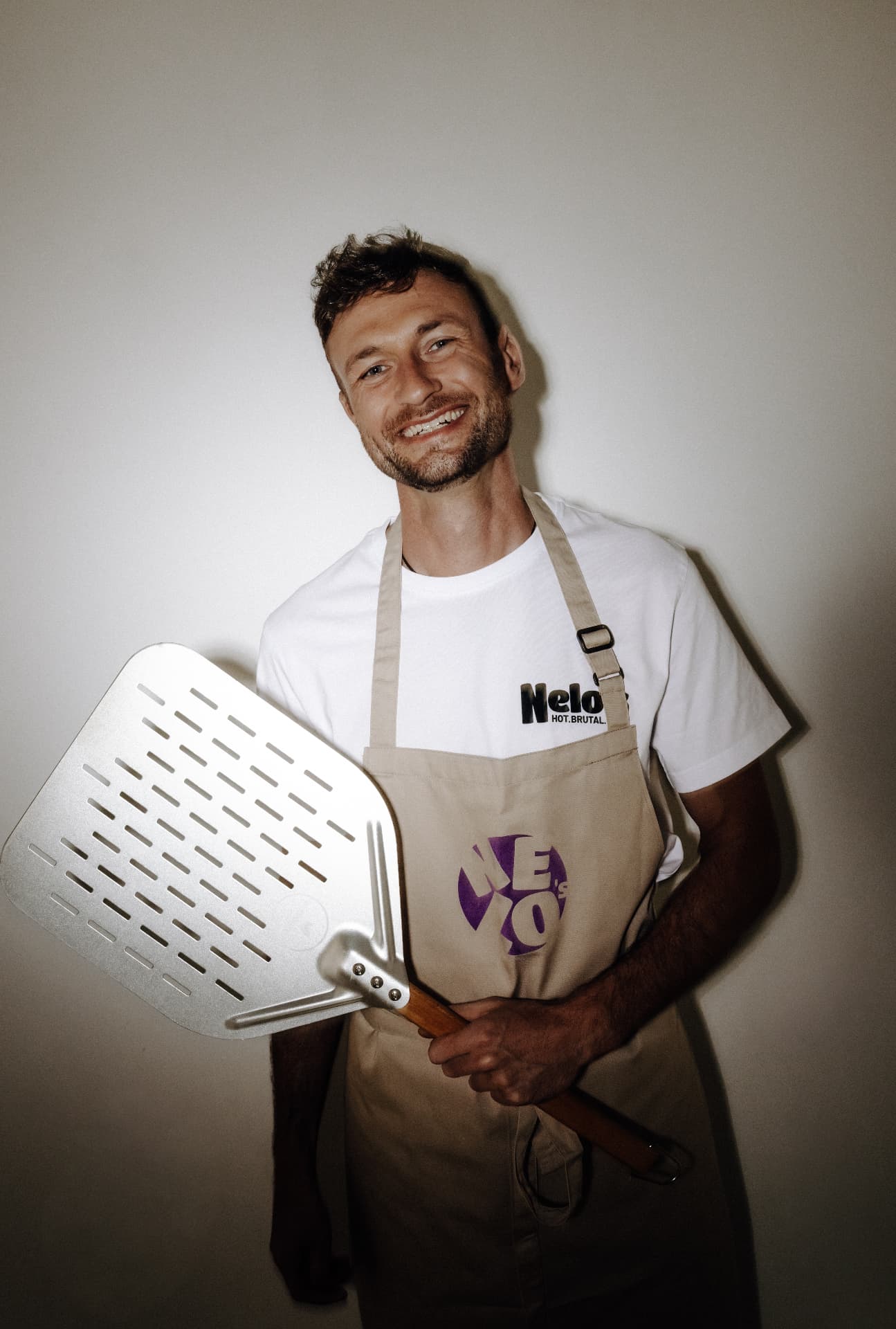 Manu von Nelo's in Schürze und Nelo's-Shirt hält eine Pizzaschaufel vor einer hellen Wand